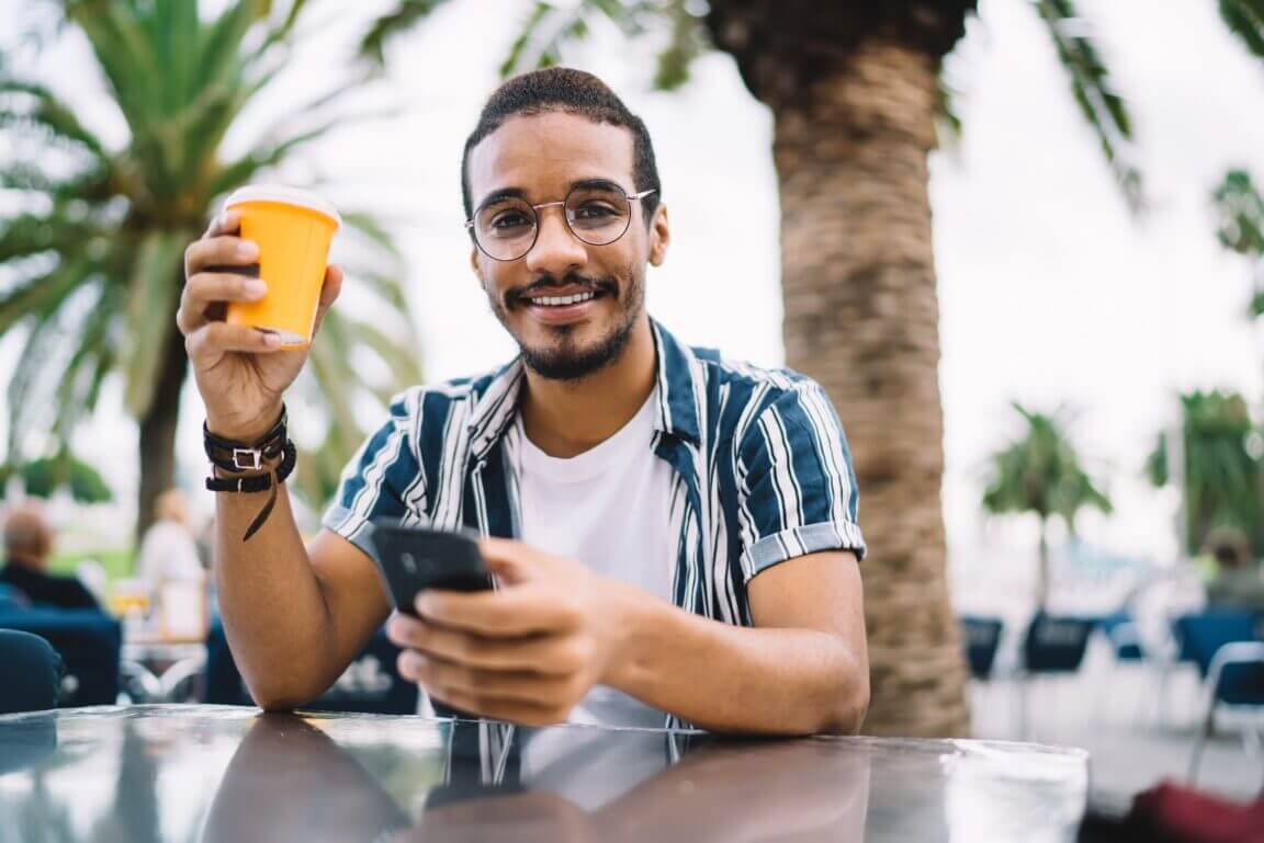 man smiles holding up a to-go coffee cup and phone