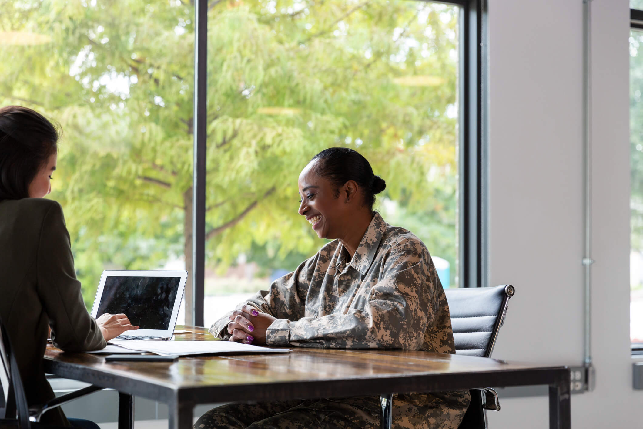 A military member and a civilian smile while working together at a table with a laptop and papers.